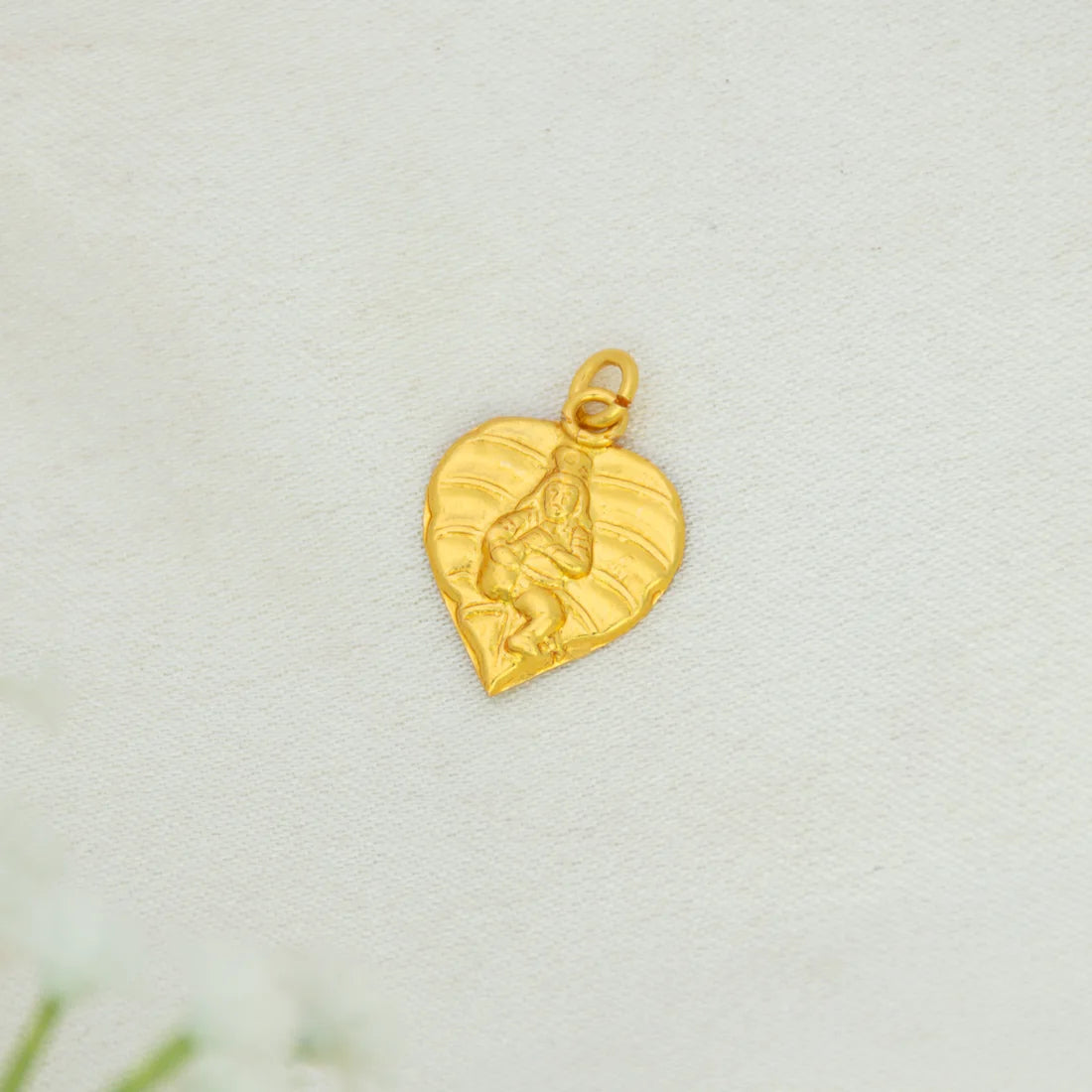 A Gold Plated Traditional Baby Krishna Alila Pendant with an embossed figure, displayed on a textured off-white surface.
