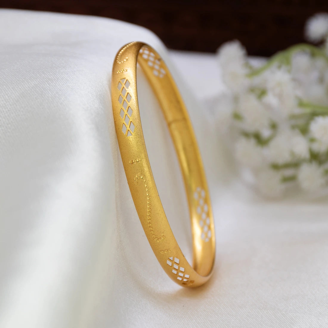 A Matte Gold-Plated Geometric Lattice Bangle with geometric cutout patterns, resting on a white textured fabric surface with a soft-focus floral background.