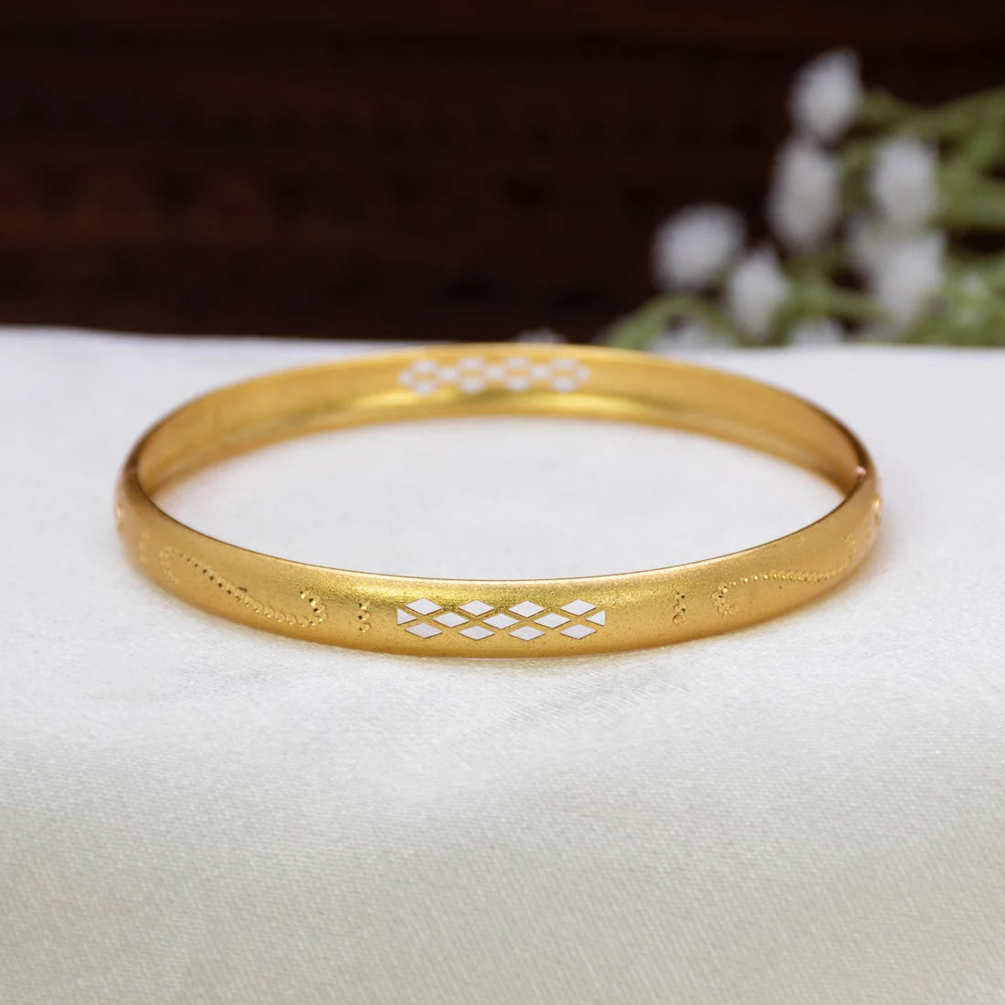 A Matte Gold-Plated Geometric Lattice Bangle with white enamel geometric patterns, resting on a white textured fabric surface with a blurred dark brown background and white flowers.