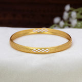 A Matte Gold-Plated Geometric Lattice Bangle with white enamel geometric patterns, resting on a white textured fabric surface with a blurred dark brown background and white flowers.