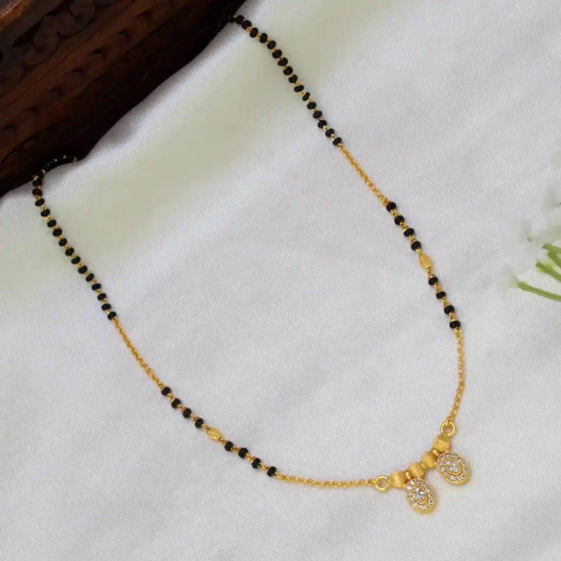 A gold and black beaded necklace with two small oval pendant charms, displayed on a white fabric surface next to a dark wooden prop.
