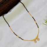 A gold and black beaded necklace with two small oval pendant charms, displayed on a white fabric surface next to a dark wooden prop.