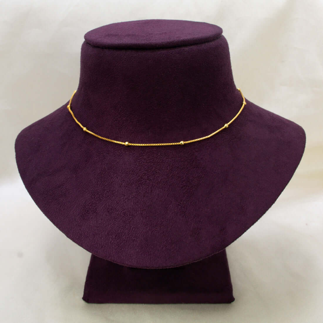 A short gold-plated beaded box chain - 15 Inches with small bead-like links displayed on a dark purple velvet jewelry bust against a light beige background.