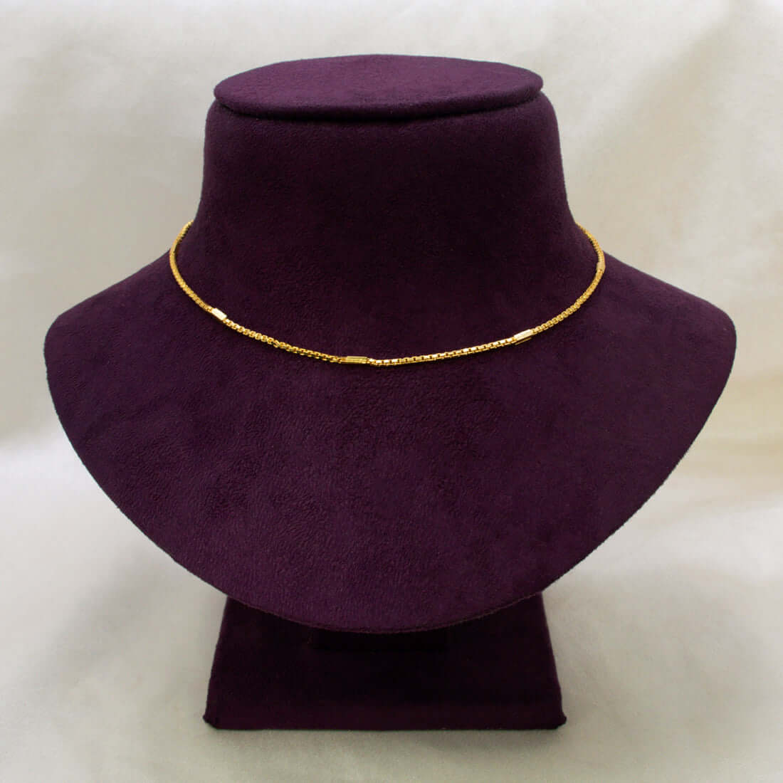 A short gold-plated designer box chain necklace displayed on a dark purple velvet jewelry bust against a light beige background.
