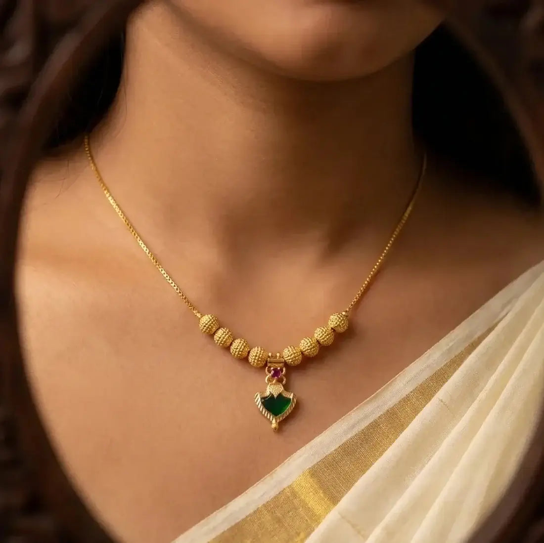 A close-up of a woman wearing a gold plated necklace with textured beads and a green Palakka-shaped pendant, styled with a cream and gold sari.