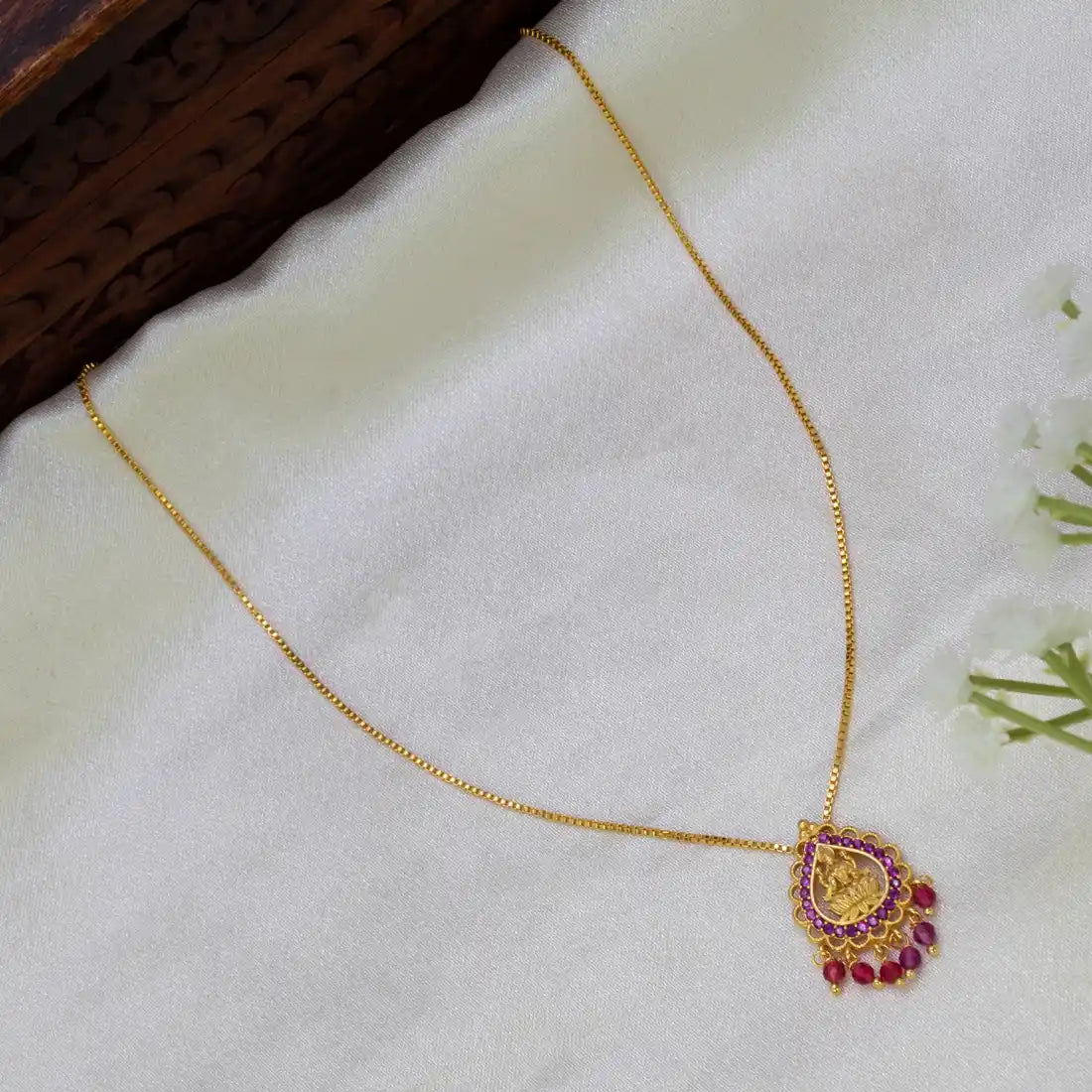 A gold plated chain necklace with a decorative pendant featuring a gold-toned  Lakshmi central motif and ruby stones, displayed on a soft white fabric surface.