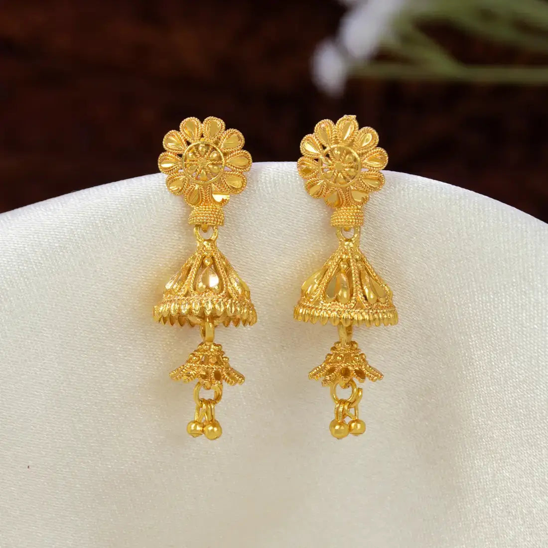A pair of ornate South Indian Gold-Plated Floral Stud Two-Step Jhumkas displayed on a white fabric stand against a dark brown blurred background.