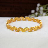 A gold-toned twisted Gold-Plated Designer Leaf Bangle, displayed on a white surface with a blurred dark brown background and small white flowers.