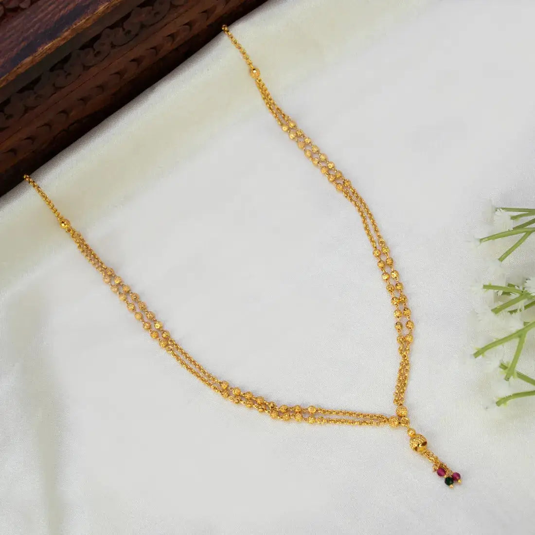 A gold chain necklace with a delicate double-strand design and a small hanging charm featuring red and green stones, displayed on a white satin fabric with a dark wooden block in the corner.