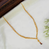 A gold chain necklace with a delicate double-strand design and a small hanging charm featuring red and green stones, displayed on a white satin fabric with a dark wooden block in the corner.