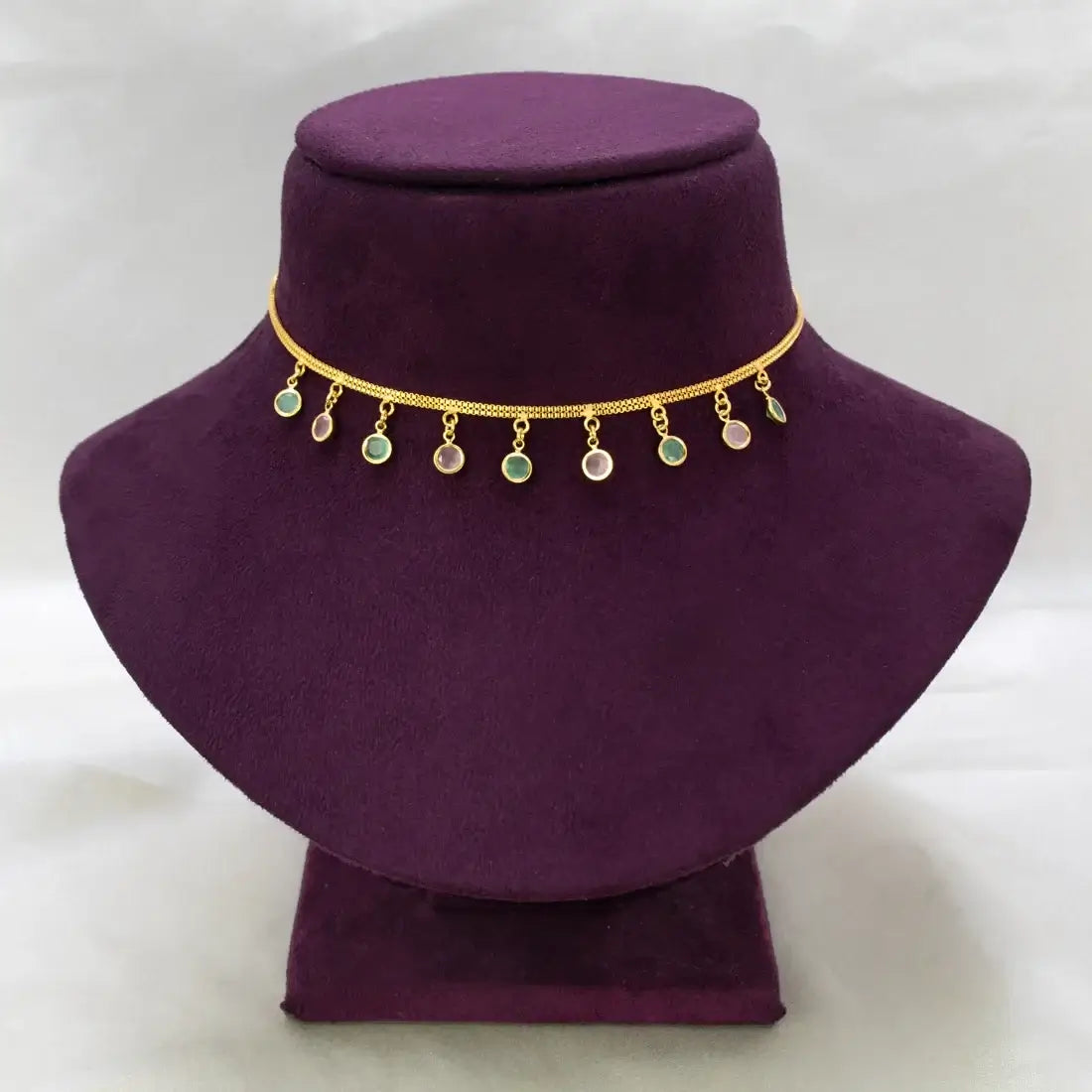 A gold chain necklace with small dangling round charms, including green and iridescent stones, displayed on a purple velvet jewelry bust against a white background.