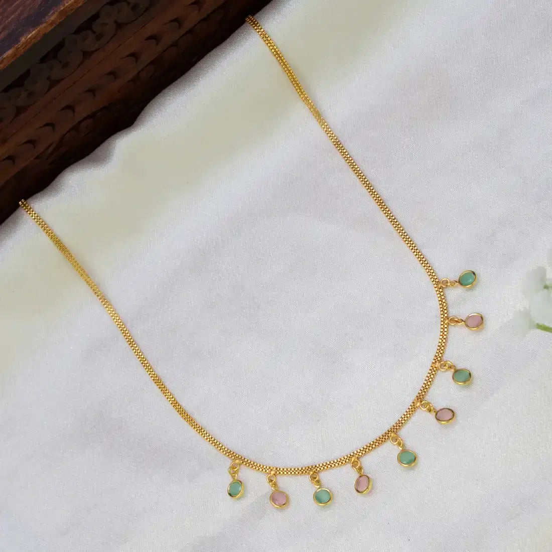 A gold chain necklace with seven small round dangling charms, each featuring a light green and pink iridescent stone, displayed on a white textured fabric surface.