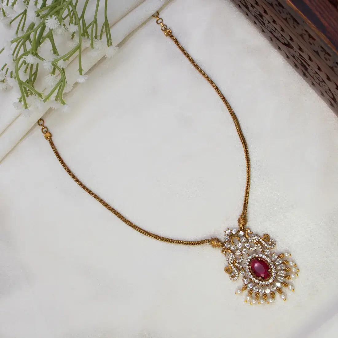 Victorian Antique Gold Peacock Stone Pendant Necklace with an ornate pendant featuring a central oval red gemstone and surrounding clear stones, displayed on a white fabric surface with small white flowers in the corner.