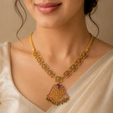 A close-up of a woman wearing a gold-toned traditional necklace with a fan-shaped pendant, decorated with pink and green stones.