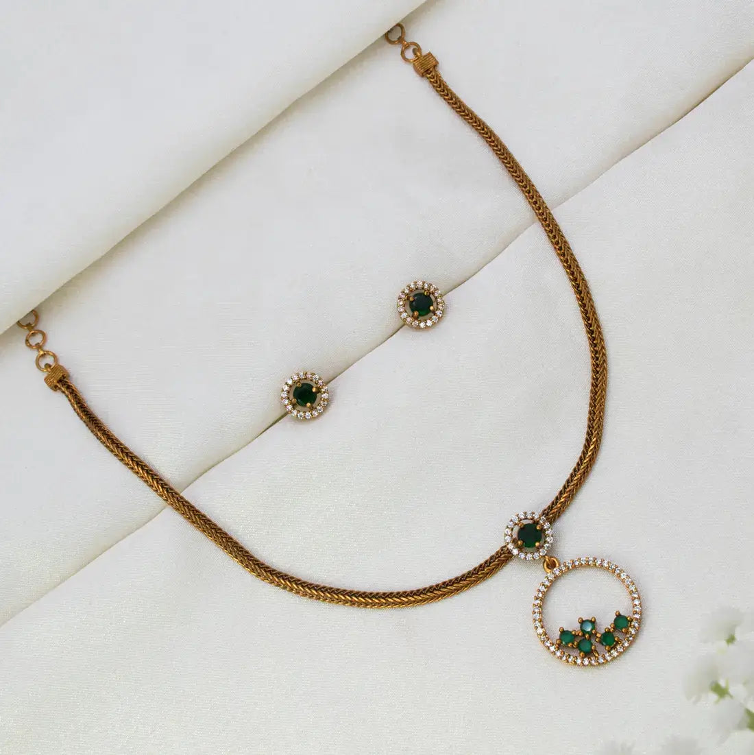A Victorian Gold-Plated Stone Necklace & Earring Set displayed on a soft white fabric background. The necklace has a textured chain, a small oval charm with a green stone surrounded by clear stones, and a larger circular pendant featuring a curved row of green stones bordered by clear stones.