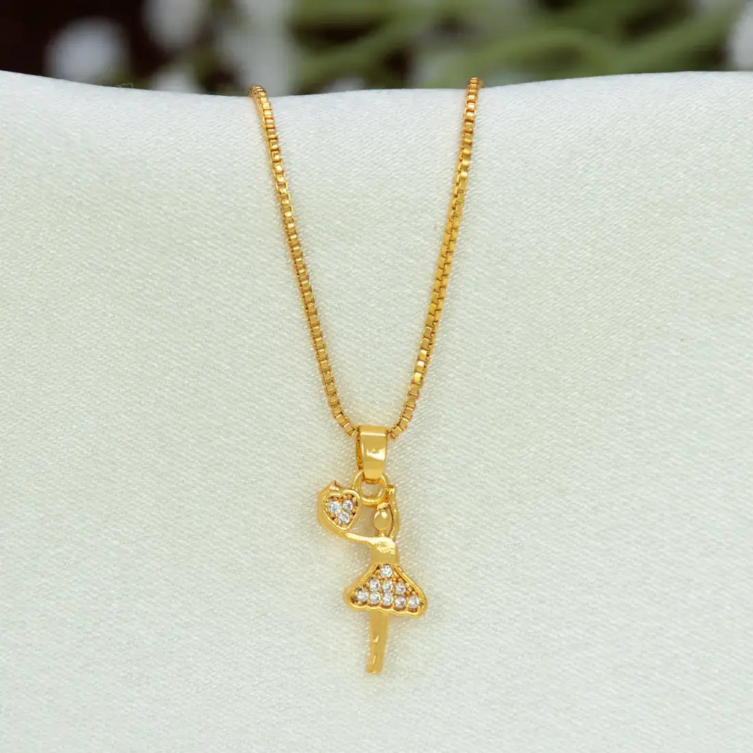 A gold chain with a gold-plated ballerina dancer pendant and a small heart-shaped charm, displayed on a white jewelry bust.